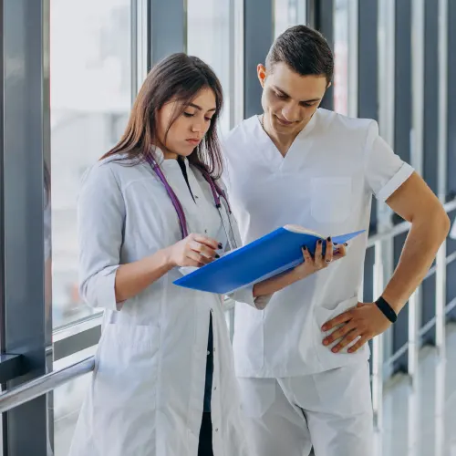 Eine brünette Ärztin im weißen Kittel mit Stethoskop um den Halsund ein braunhaariger Pfleger in weißem T-Shirt und Hose stehen nebeneinander und lesen in einer blauen Mappe