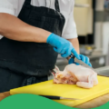 Eine Person mit schwarzer Schürze und blauen Einmalhandschuhen schneidet mit einem Messer ein Hühnchen auf einem gelben Schneidbrett
