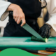 Eine Person in Kochjacke mit schwarzer Schürze und einem schwarzen Einmalhandschuh schneidet eine Gurke auf einem grünen Schneidbrett