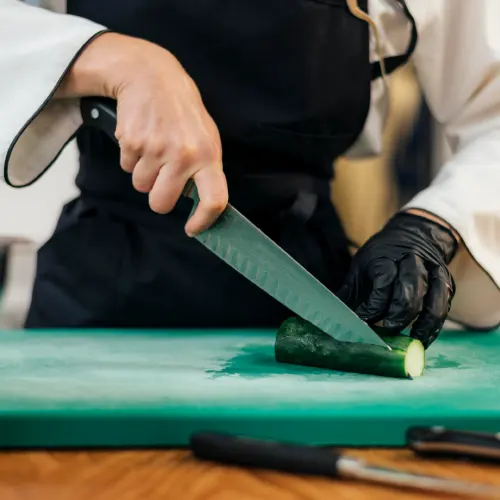 Eine Person in Kochjacke mit schwarzer Schürze und einem schwarzen Einmalhandschuh schneidet eine Gurke auf einem grünen Schneidbrett