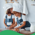 Zwei Kinder mit Kochmützen und Jeansschürzen backen mit Eiern und Mehl
