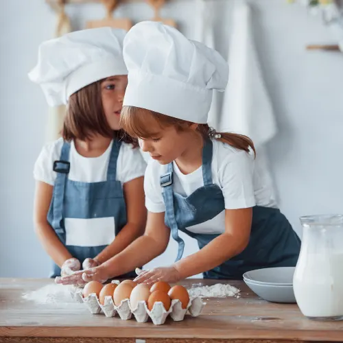 Zwei Kinder mit Kochmützen und Jeansschürzen backen mit Eiern und Mehl
