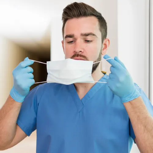 Braunhaariger Mann in blauem Pflegekittel und mit blauen Einmalhandschuhen zieht sich gerade seinen weißen Mundschutz an